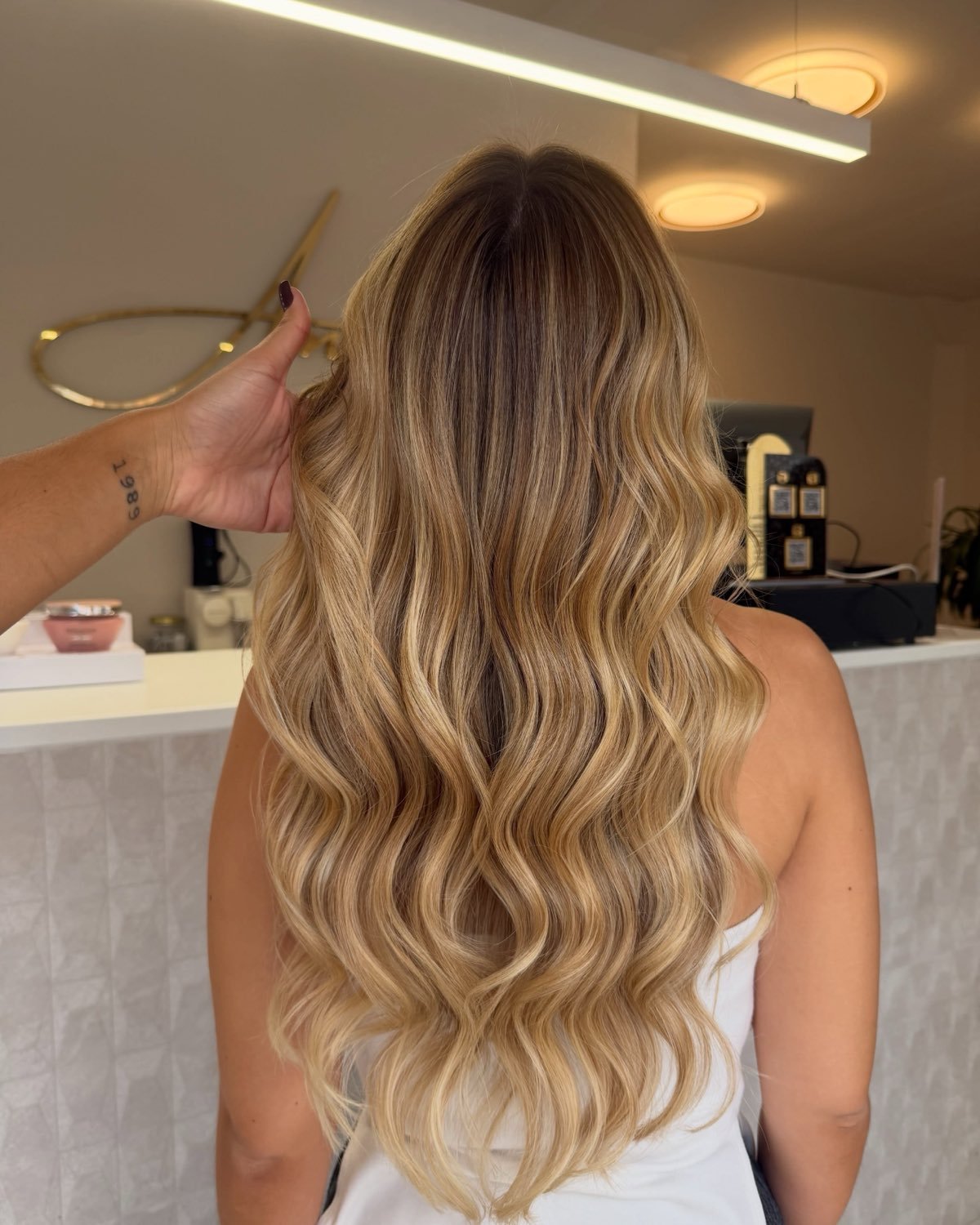 Cabelo loiro dourado com ondas largas, visto de costas, num salão claro com luzes redondas no teto