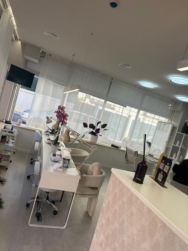 Zona de manicure com mesas brancas, cadeiras claras e amplas janelas com luz natural.