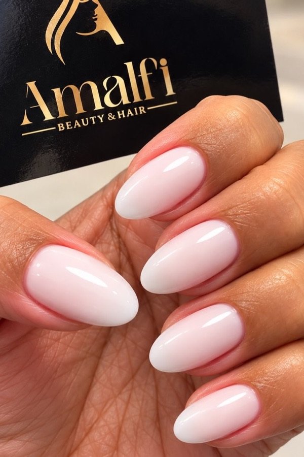 Unhas amendoadas em gel rosa leitoso, com um cartão Amalfi ao fundo